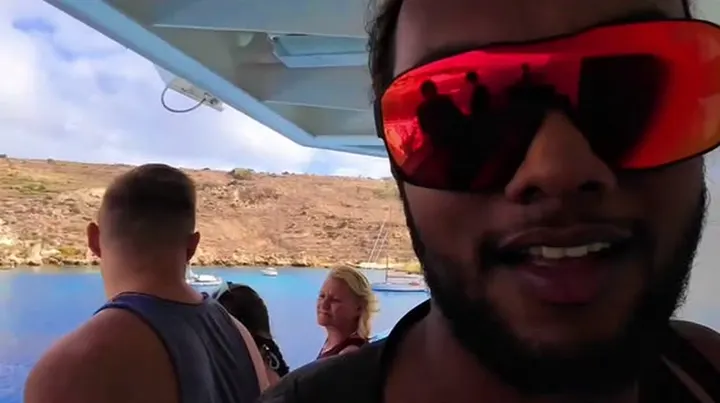 Ray grinning at the camera in red sunglasses on a ferry, coastline behind him